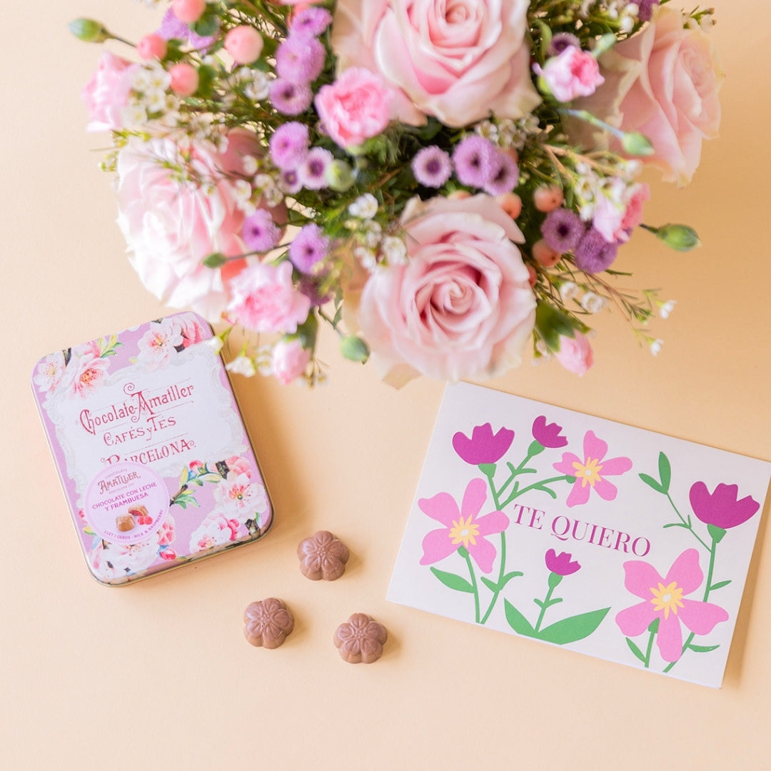 Detalle del arreglo de flores frescas en tonos rosas Dulce base estriada verde menta, junto a una lata vintage de bombones de chocolate Amatller Barcelona y una tarjeta regalo ilustrada con la frase "TE QUIERO" sobre un fondo liso de color crema.