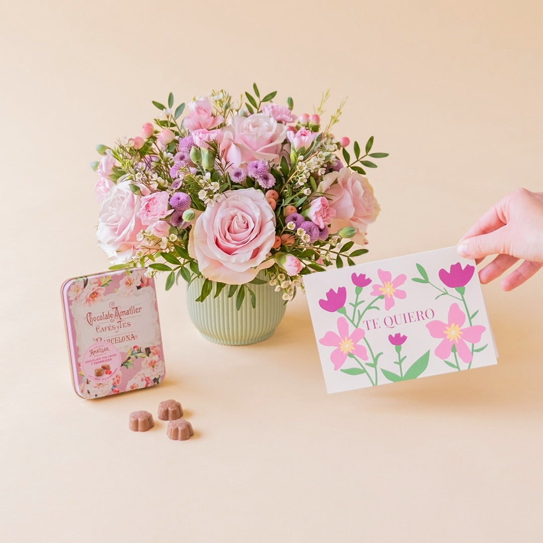 Arreglo de flores frescas en tonos rosas Dulce base estriada verde menta, junto a una lata vintage de bombones de chocolate Amatller Barcelona y una mano sosteniendo una tarjeta regalo ilustrada con la frase "TE QUIERO" sobre un fondo liso de color crema.