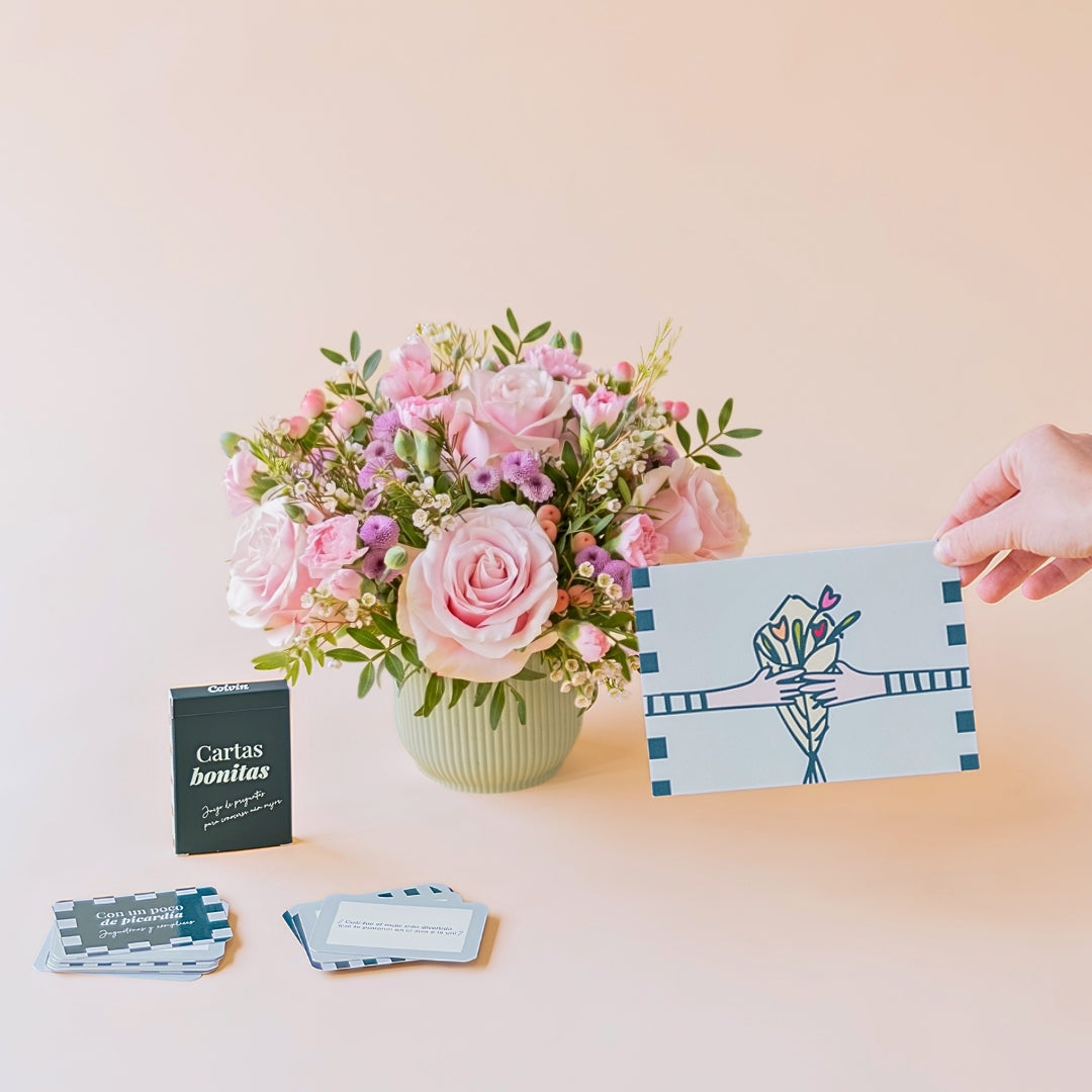 Arreglo de flores frescas en tonos rosas Dulce con base verde menta, junto a la caja verde oscuro del juego de mesa "Cartas bonitas" de Colvin con cartas de preguntas desplegadas y una mano sosteniendo una tarjeta de felicitación ilustrada sobre un fondo liso de color crema.