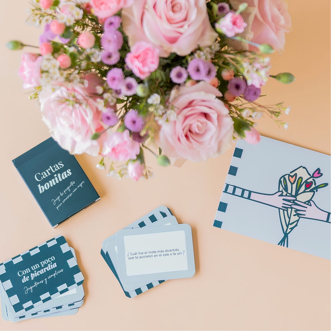 Detalle del arreglo de flores frescas en tonos rosas Dulce con base verde menta, junto a la caja verde oscuro del juego de mesa "Cartas bonitas" de Colvin con cartas de preguntas desplegadas y una tarjeta de felicitación ilustrada sobre un fondo liso de color crema.