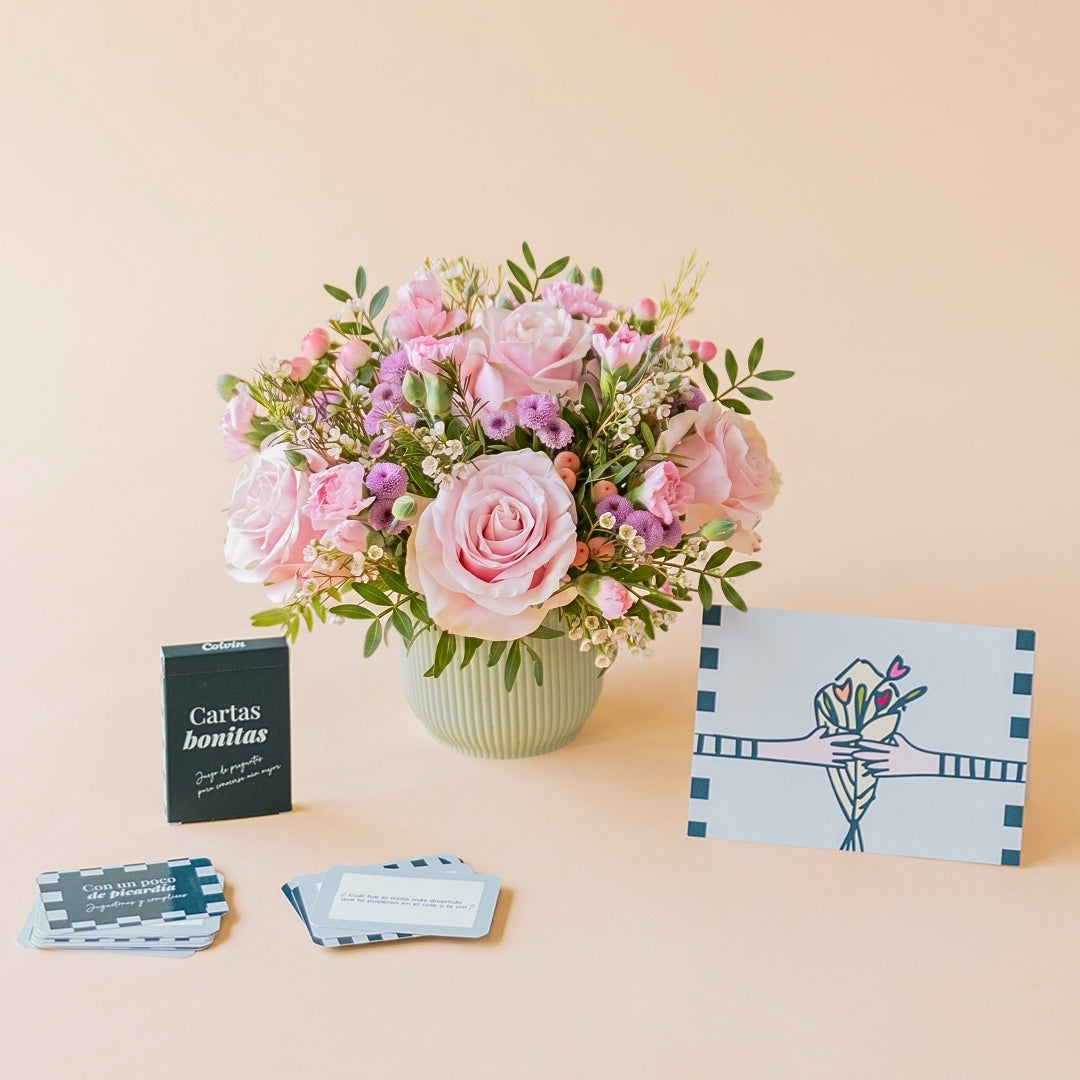 Arreglo de flores frescas en tonos rosas Dulce con base verde menta, junto a la caja verde oscuro del juego de mesa "Cartas bonitas" de Colvin con cartas de preguntas desplegadas y una tarjeta de felicitación ilustrada sobre un fondo liso de color crema.