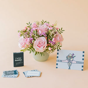 Arreglo de flores frescas en tonos rosas Dulce con base verde menta, junto a la caja verde oscuro del juego de mesa "Cartas bonitas" de Colvin con cartas de preguntas desplegadas y una tarjeta de felicitación ilustrada sobre un fondo liso de color crema.