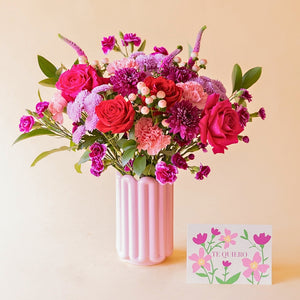 Ramo El Hechizo de Colvin con rosas rojas y fucsia, crisantemos lila y claveles en jarrón Lola rosa palo, junto a tarjeta floral "Te Quiero"