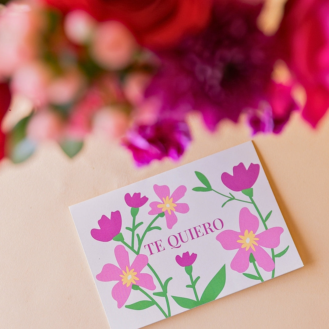 Primer plano de tarjeta Más que Flores "Te Quiero" con ilustración floral rosa y lila, con ramo de rosas rojas y fucsia de Colvin al fondo