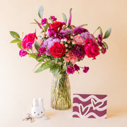 Ramo El Hechizo de Colvin con rosas rojas y fucsia, crisantemos lila y claveles en jarrón de cristal, junto a llavero Miffy y tarjeta de corazones burdeos