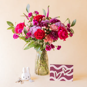 Ramo El Hechizo de Colvin con rosas rojas y fucsia, crisantemos lila y claveles en jarrón de cristal, junto a llavero Miffy y tarjeta de corazones burdeos