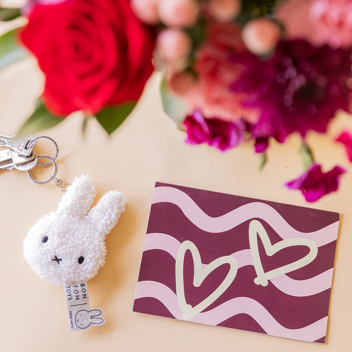 Detalle de llavero Miffy Bon Ton Toys y tarjeta de corazones burdeos y rosa, con rosas rojas y claveles fucsia de Colvin al fondo