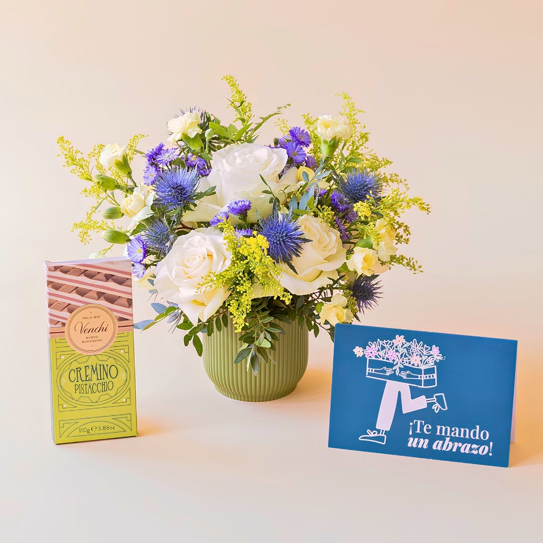 Arreglo de flores frescas El Mágico de Colvin con rosas blancas premium, cardos azules y detalles silvestres en una base estriada de verde oliva, junto a una tableta de chocolate Venchi Cremino Pistacchio y una mano sosteniendo una tarjeta de felicitación azul con la frase "¡Te mando un abrazo!" sobre un fondo liso de color crema.