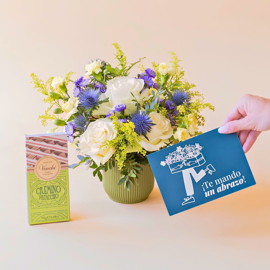 Arreglo de flores frescas El Mágico de Colvin con rosas blancas premium, cardos azules y detalles silvestres en una base estriada de verde oliva, junto a una tableta de chocolate Venchi Cremino Pistacchio y una mano sosteniendo una tarjeta de felicitación azul con la frase "¡Te mando un abrazo!" sobre un fondo liso de color crema.