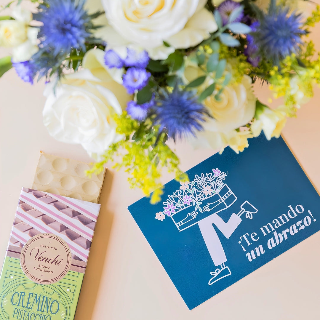 Detalle del arreglo de flores frescas El Mágico de Colvin con rosas blancas premium, cardos azules y detalles silvestres, junto a una tableta de chocolate Venchi Cremino Pistacchio y una tarjeta de felicitación azul con la frase "¡Te mando un abrazo!" sobre un fondo liso de color crema.