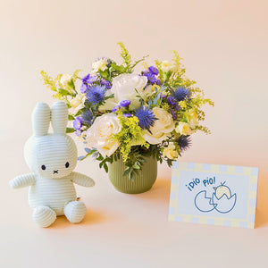 Arreglo de flores frescas El Mágico de Colvin con rosas blancas premium, cardos azules y detalles silvestres en una base estriada verde oliva, junto a un peluche Miffy en pana color verde menta y una tarjeta de nacimiento ilustrada con un pollito y la frase "¡Pío pío!" sobre un fondo crema.