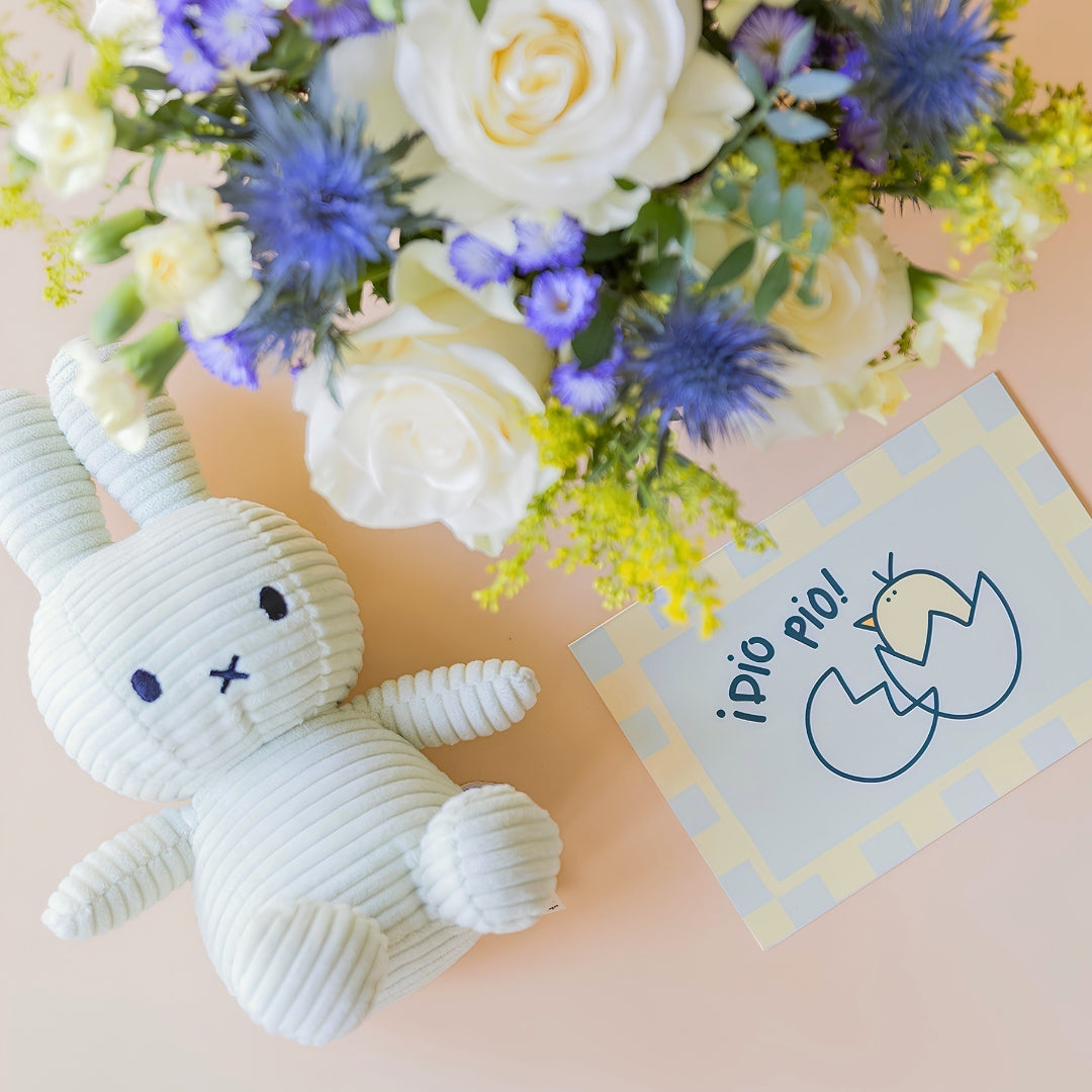 Detalle del arreglo de flores frescas El Mágico de Colvin con rosas blancas premium, cardos azules, junto a un peluche Miffy en pana color verde menta y una tarjeta de nacimiento ilustrada con un pollito y la frase "¡Pío pío!" sobre un fondo crema.