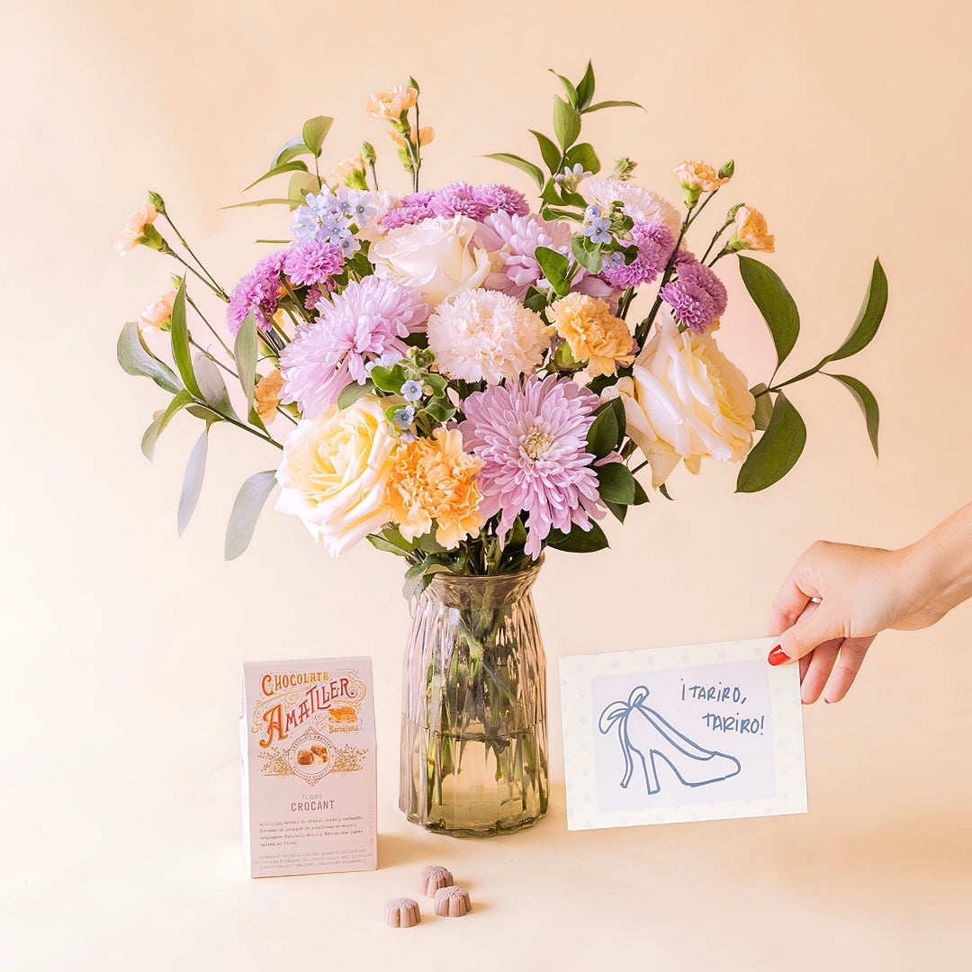 Mano sosteniendo tarjeta "¡Tariro, Tariro!" junto a ramo de flores pastel de Colvin en jarrón de cristal con bombones Amatller Crocant
