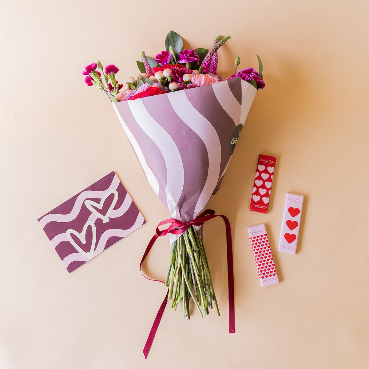 Ramo El Hechizo de Colvin con rosas rojas, crisantemos lila e hipericum blanco en envoltorio de ondas, junto a tarjeta de corazones y chocolatinas Simón Coll