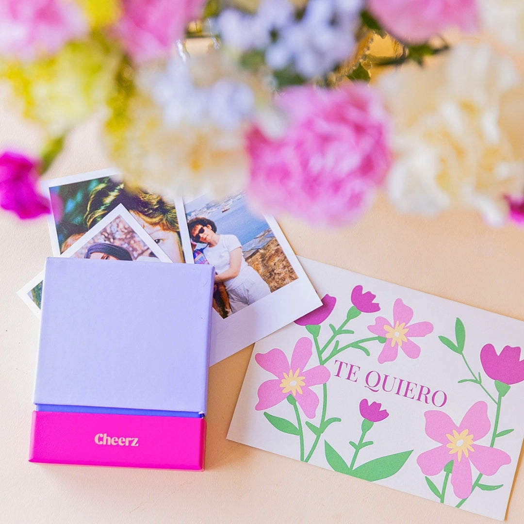 Cheerz Box lila y fucsia abierta con fotos reveladas en formato instax, junto a tarjeta "Te Quiero" con flores ilustradas, con ramo de claveles pastel desenfocado al fondo