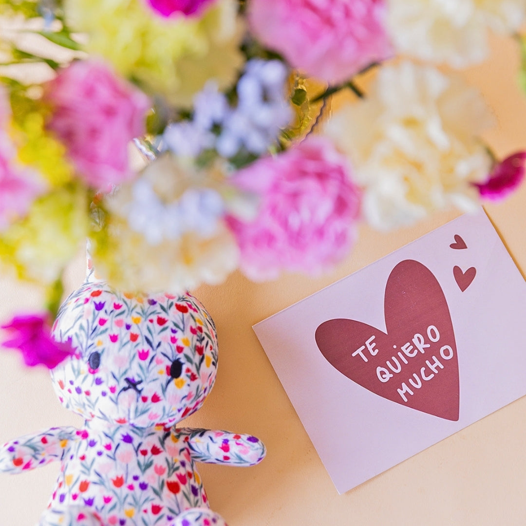 Primer plano de peluche Miffy de tela floral y tarjeta Te Quiero Mucho con corazón rosa, con ramo de claveles crema, rosa y fucsia de Colvin desenfocado al fondo