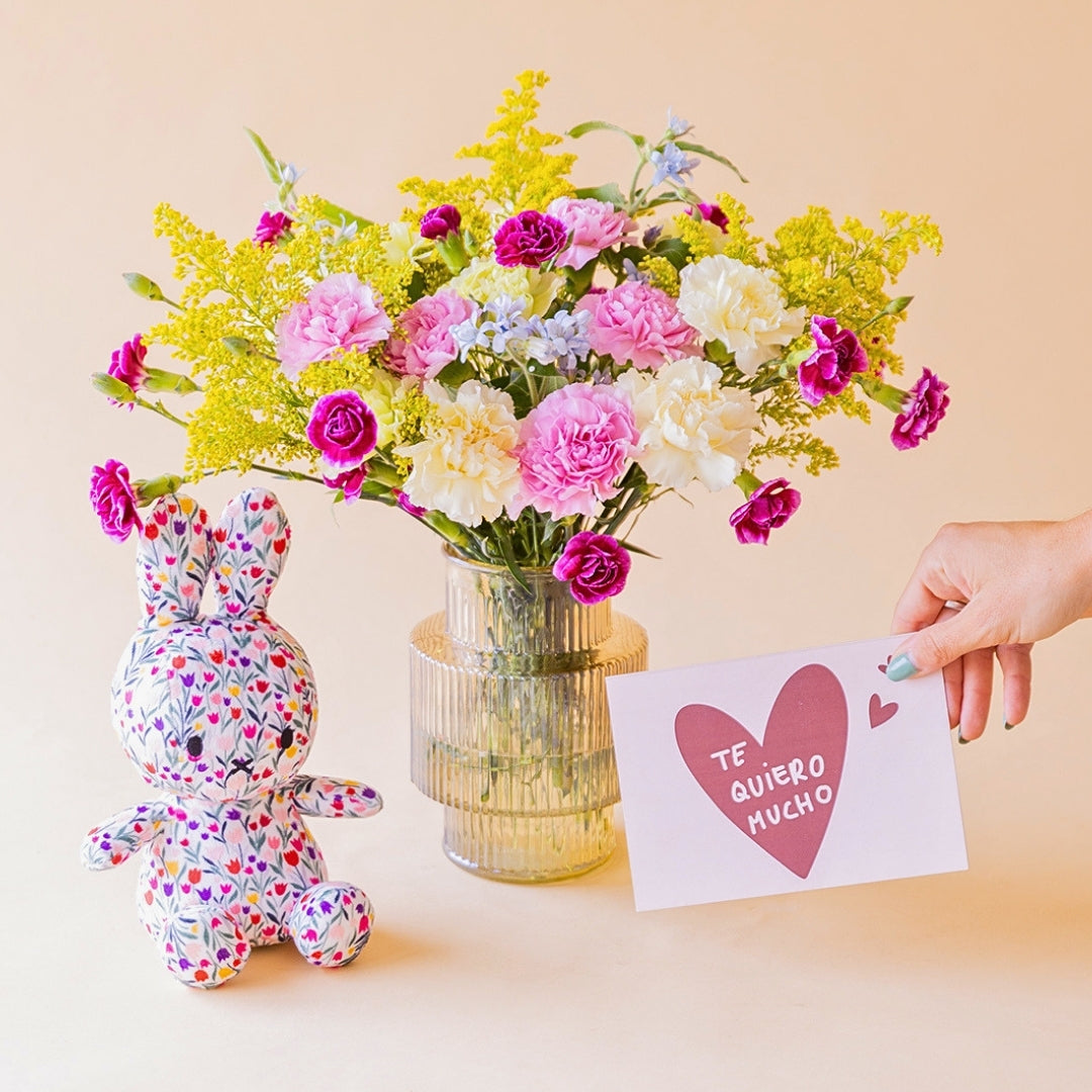 Ramo de claveles crema, rosa, fucsia y solidago amarillo de Colvin en jarrón acanalado transparente, con peluche Miffy de tela floral y mano sosteniendo tarjeta Te Quiero Mucho, sobre fondo melocotón