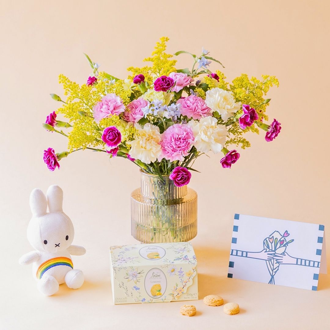 Ramo de claveles crema, rosas, fucsia y solidago amarillo de Colvin en jarrón acanalado transparente, con peluche Miffy arcoíris, caja de galletas Loison Limone y tarjeta ilustrada, sobre fondo melocotón