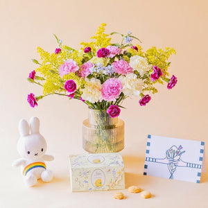 Ramo de claveles crema, rosas, fucsia y solidago amarillo de Colvin en jarrón acanalado transparente, con peluche Miffy arcoíris, caja de galletas Loison Limone y tarjeta ilustrada, sobre fondo melocotón
