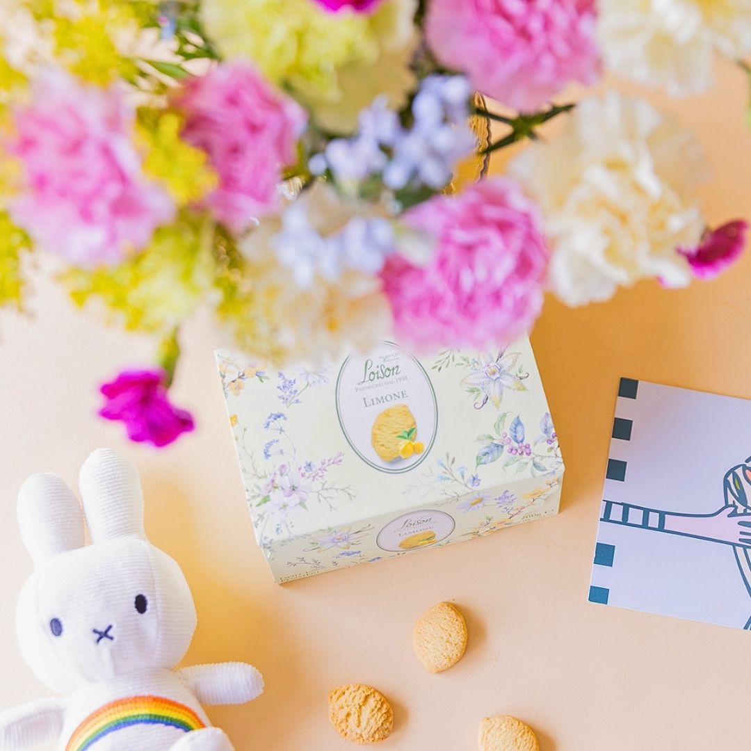 Detalle de caja de galletas Loison Limone con peluche Miffy arcoíris y tarjeta ilustrada, junto al ramo de claveles crema y rosas de Colvin al fondo