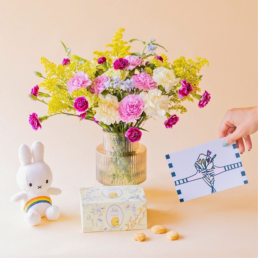 Ramo de claveles crema, rosas y fucsia de Colvin en jarrón acanalado transparente con peluche Miffy arcoíris, caja de galletas Loison y mano sosteniendo tarjeta ilustrada, sobre fondo melocotón