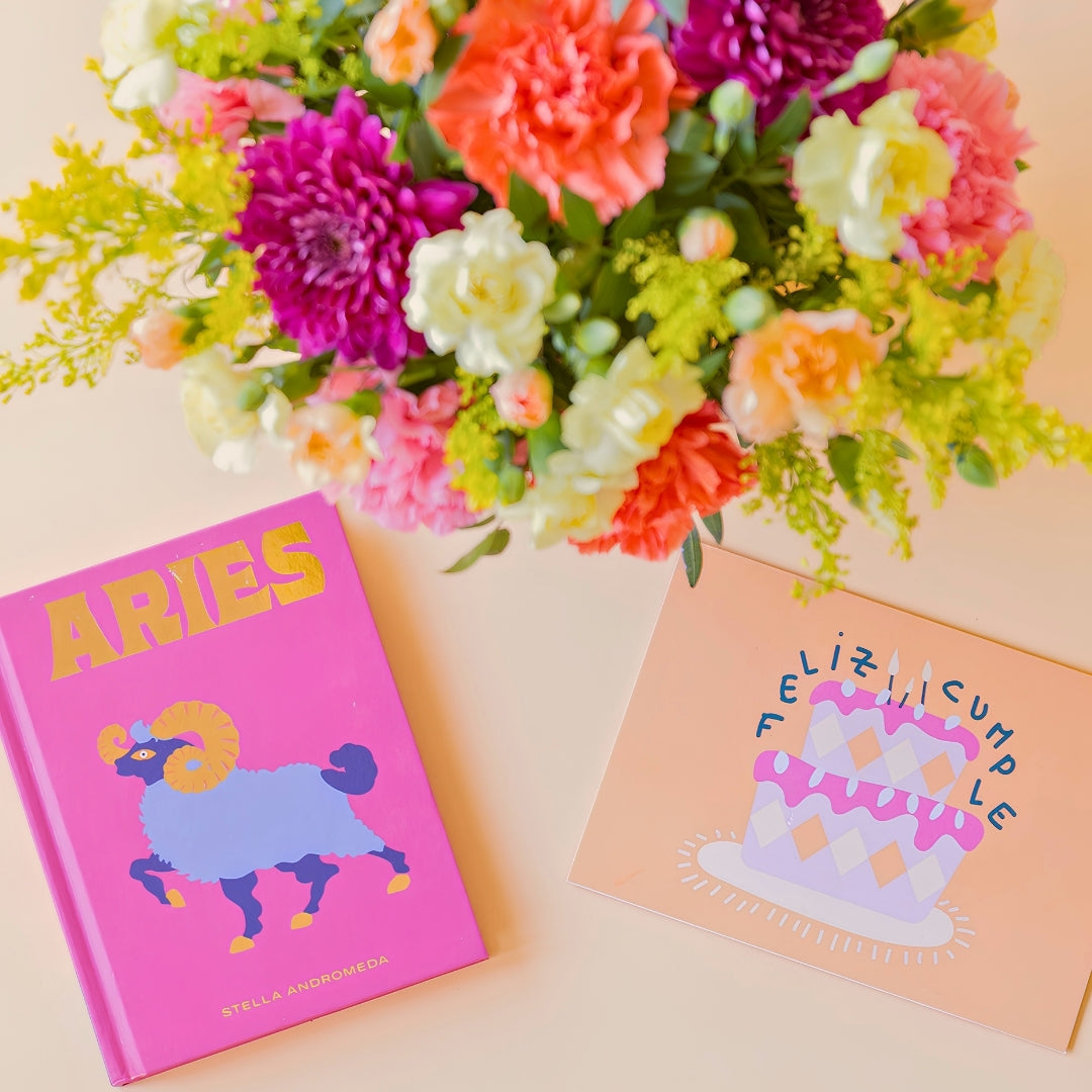 Detalle del arreglo de flores frescas multicolores La Chispa de Colvin con claveles en tonos rosas y melocotón y crisantemos morados, junto al libro del horóscopo Aries de tapa dura con portada rosa y una tarjeta de cumpleaños de Feliz Cumple sobre un fondo crema.