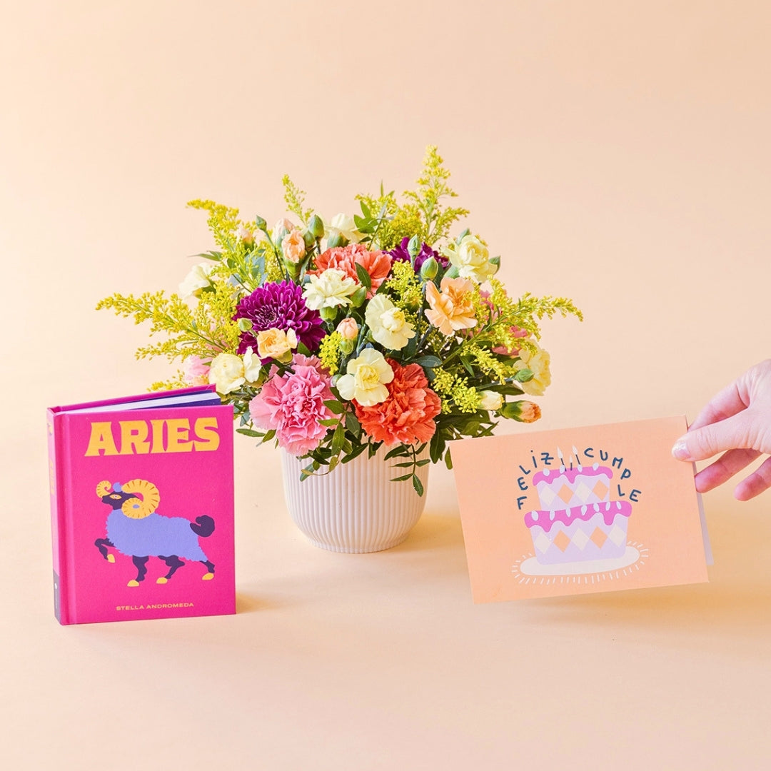 Arreglo de flores frescas multicolores La Chispa de Colvin con claveles en tonos rosas y melocotón y crisantemos morados en una base estriada blanca, junto al libro del horóscopo Aries de tapa dura con portada rosa y una mano sosteniendo una tarjeta de cumpleaños de Feliz Cumple sobre un fondo crema.