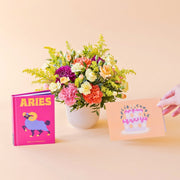 Arreglo de flores frescas multicolores La Chispa de Colvin con claveles en tonos rosas y melocotón y crisantemos morados en una base estriada blanca, junto al libro del horóscopo Aries de tapa dura con portada rosa y una mano sosteniendo una tarjeta de cumpleaños de Feliz Cumple sobre un fondo crema.