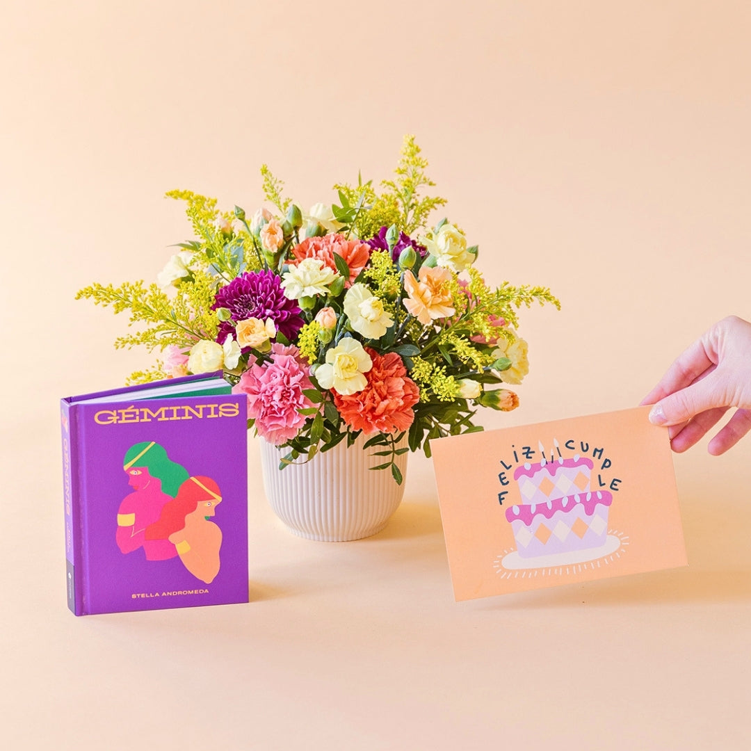 Arreglo de flores frescas multicolores La Chispa de Colvin con claveles en tonos rosas y melocotón y crisantemos morados en una base estriada blanca, junto al libro del horóscopo Géminis de tapa dura con portada morada y una mano sosteniendo una tarjeta de cumpleaños de Feliz Cumple sobre un fondo crema.