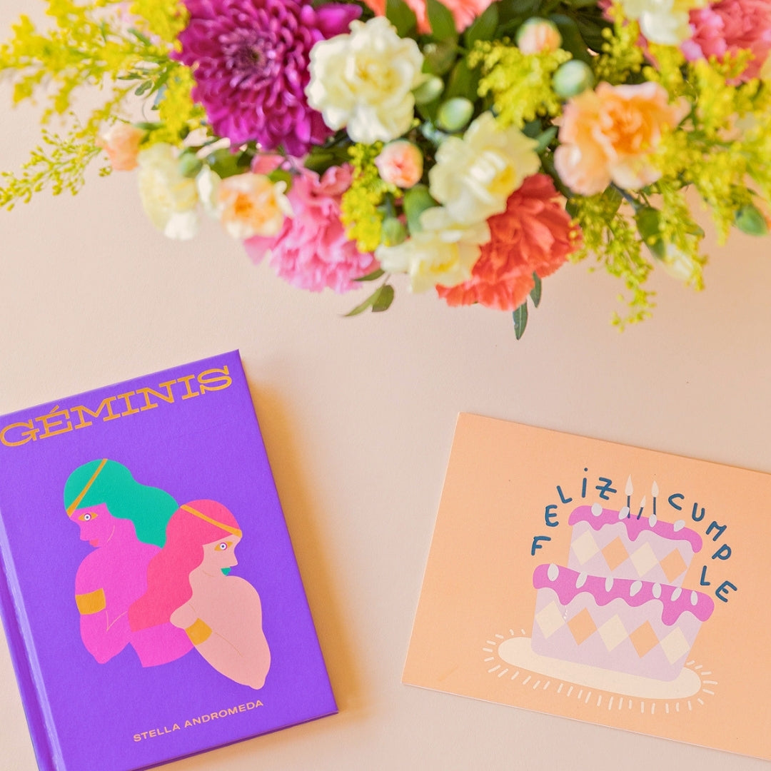 Detalle del arreglo de flores frescas multicolores La Chispa de Colvin con claveles en tonos rosas y melocotón y crisantemos morados, junto al libro del horóscopo Géminis de tapa dura con portada morada y una tarjeta de cumpleaños de Feliz Cumple sobre un fondo crema.