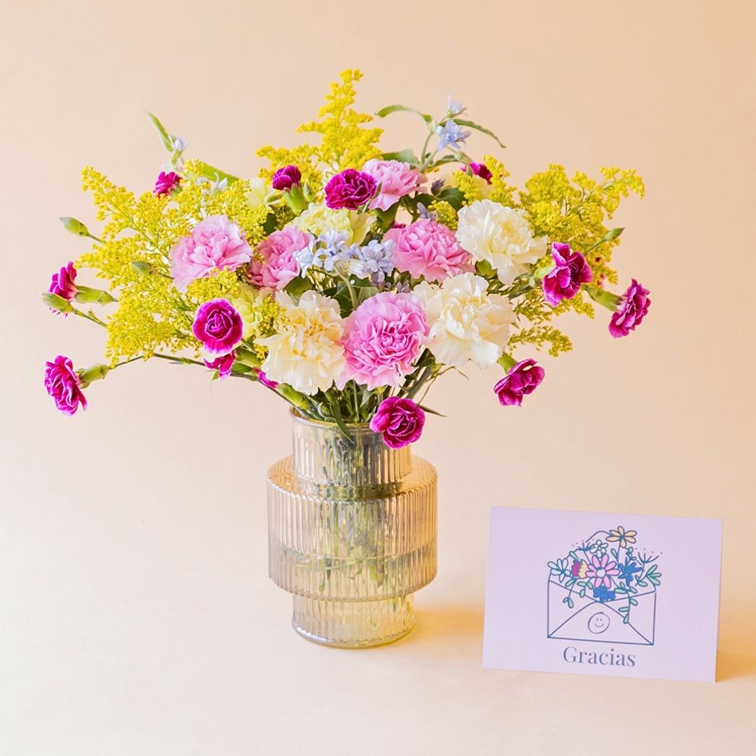 Ramo La Alegría de Colvin con claveles fucsia, rosa, crema y solidago amarillo en jarrón acanalado dorado, junto a tarjeta Gracias, sobre fondo melocotón