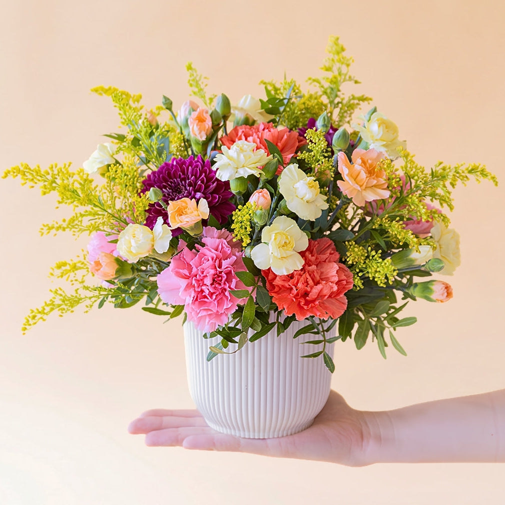 Mano sosteniendo el La Chispa - Detalle de arreglo floral con claveles amarillos y naranjas con crisantemos morados de Colvin.
