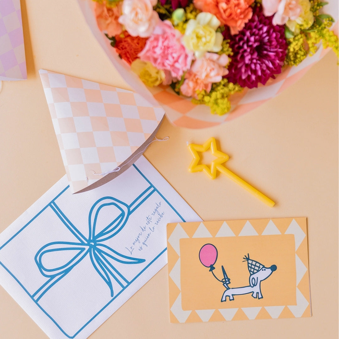 Detalle de arreglo de flores frescas coloridas La Chispa de Colvin, gorros de fiesta a juego, una vela pequeña de estrella amarilla y una tarjeta de cumpleaños ilustrada con un perro con globo sobre un fondo crema.