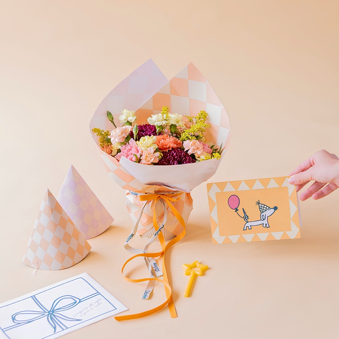 Arreglo de flores frescas coloridas La Chispa de Colvin, con claveles en tonos rosa y melocotón y crisantemos morados, envuelto en papel de diseño con patrón de cuadros color melocotón y lazo naranja, junto a dos gorros de fiesta a juego, una vela pequeña de estrella amarilla y una mano sosteniendo una tarjeta de cumpleaños ilustrada con un perro con globo sobre un fondo crema.