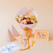 Arreglo de flores frescas coloridas La Chispa de Colvin, con claveles en tonos rosa y melocotón y crisantemos morados, envuelto en papel de diseño con patrón de cuadros color melocotón y lazo naranja, junto a dos gorros de fiesta a juego, una vela pequeña de estrella amarilla y una tarjeta de cumpleaños ilustrada con un perro con globo sobre un fondo crema.