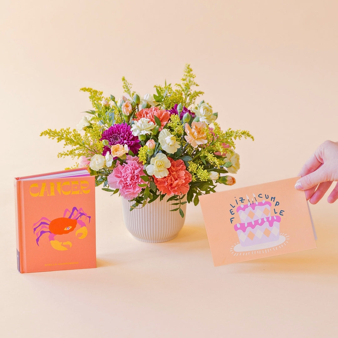 Arreglo de flores frescas multicolores La Chispa de Colvin con claveles en tonos rosas y melocotón y crisantemos morados en una base estriada blanca, junto al libro del horóscopo Cáncer de tapa dura con portada naranja y una mano sosteniendo una tarjeta de cumpleaños de Feliz Cumple sobre un fondo crema.