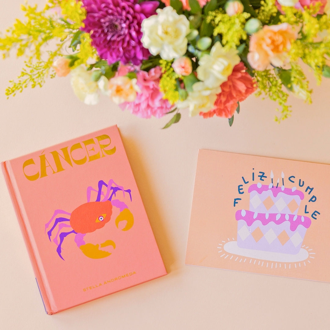Detalle del arreglo de flores frescas multicolores La Chispa de Colvin con claveles en tonos rosas y melocotón y crisantemos morados, junto al libro del horóscopo Cáncer de tapa dura con portada naranja y una tarjeta de cumpleaños de Feliz Cumple sobre un fondo crema.