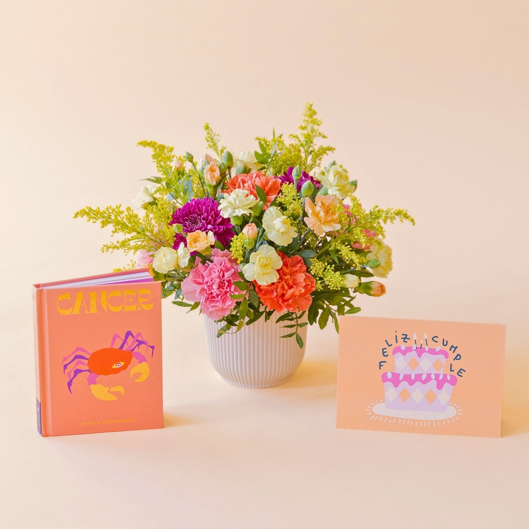 Arreglo de flores frescas multicolores La Chispa de Colvin con claveles en tonos rosas y melocotón y crisantemos morados en una base estriada blanca, junto al libro del horóscopo Cáncer de tapa dura con portada naranja y una tarjeta de cumpleaños de Feliz Cumple sobre un fondo crema.