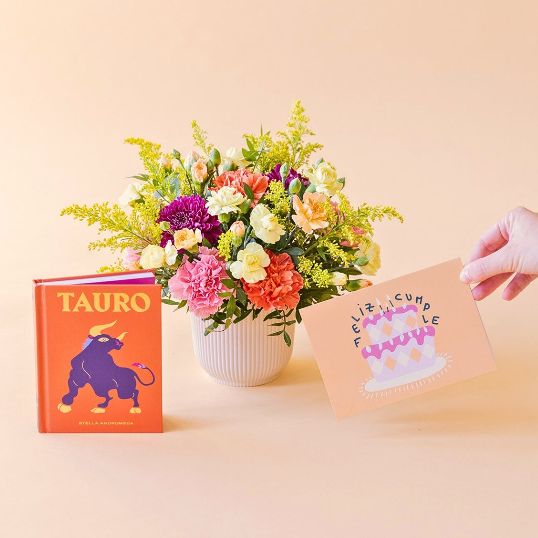 Arreglo de flores frescas multicolores La Chispa de Colvin con claveles en tonos rosas y melocotón y crisantemos morados en una base estriada blanca, junto al libro del horóscopo Tauro de tapa dura con portada naranja y una mano sosteniendo una tarjeta de cumpleaños de Feliz Cumple sobre un fondo crema.