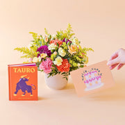 Arreglo de flores frescas multicolores La Chispa de Colvin con claveles en tonos rosas y melocotón y crisantemos morados en una base estriada blanca, junto al libro del horóscopo Tauro de tapa dura con portada naranja y una mano sosteniendo una tarjeta de cumpleaños de Feliz Cumple sobre un fondo crema.