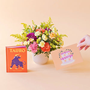 Arreglo de flores frescas multicolores La Chispa de Colvin con claveles en tonos rosas y melocotón y crisantemos morados en una base estriada blanca, junto al libro del horóscopo Tauro de tapa dura con portada naranja y una mano sosteniendo una tarjeta de cumpleaños de Feliz Cumple sobre un fondo crema.