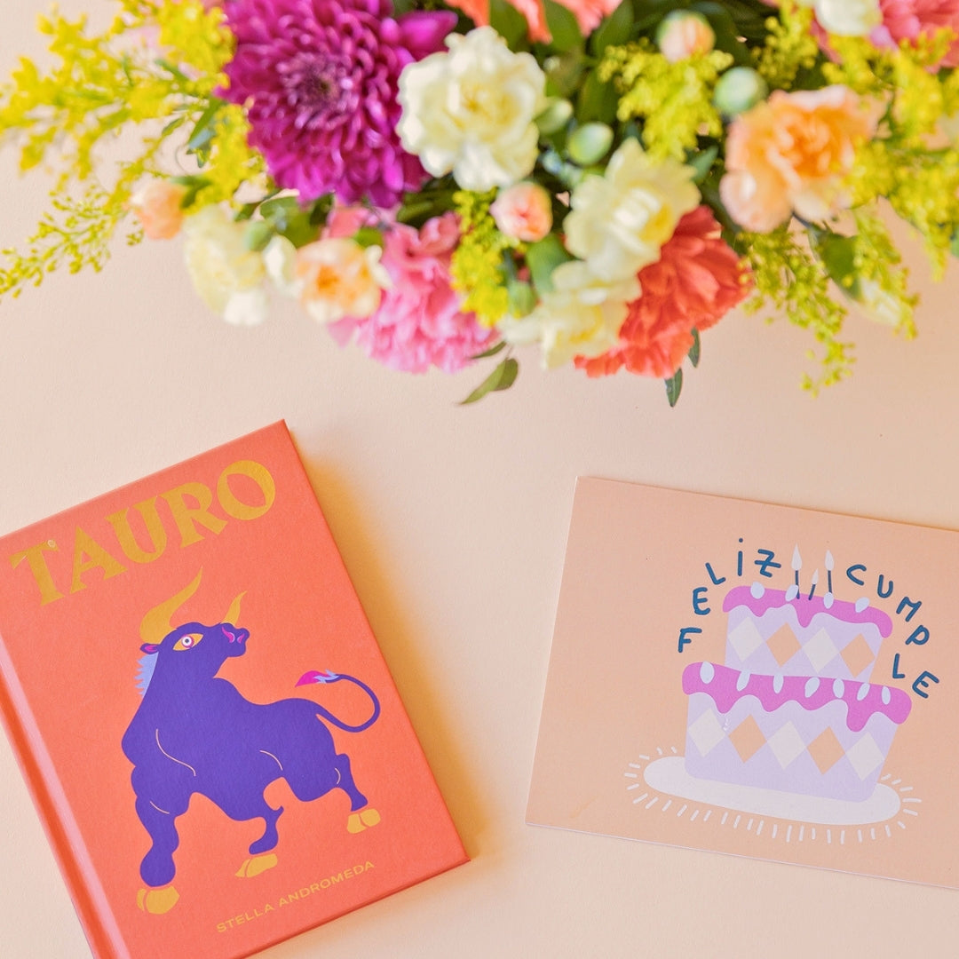 Detalle del arreglo de flores frescas multicolores La Chispa de Colvin con claveles en tonos rosas y melocotón y crisantemos morados, junto al libro del horóscopo Tauro de tapa dura con portada naranja y una mano sosteniendo una tarjeta de cumpleaños de Feliz Cumple sobre un fondo crema.