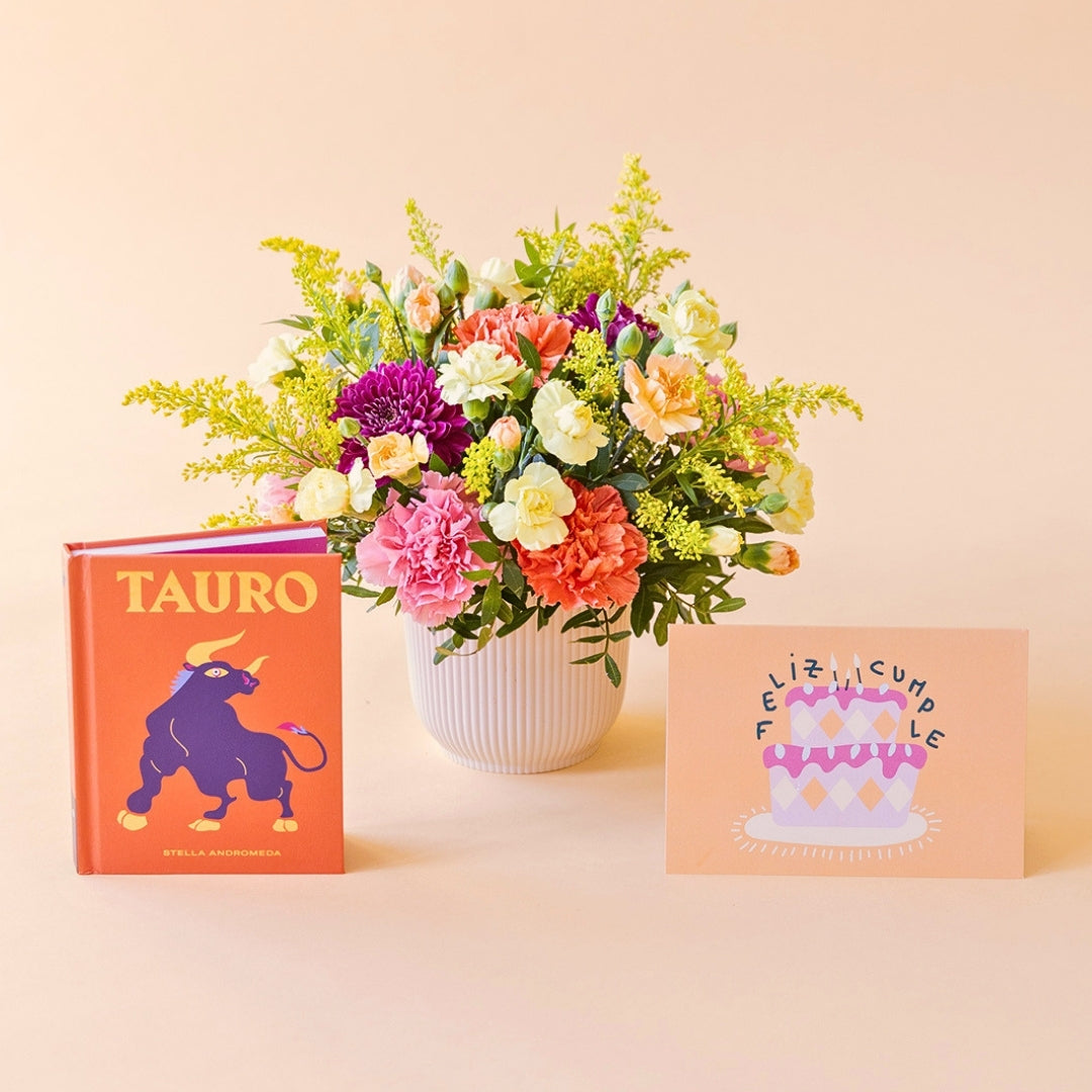 Arreglo de flores frescas multicolores La Chispa de Colvin con claveles en tonos rosas y melocotón y crisantemos morados en una base estriada blanca, junto al libro del horóscopo Tauro de tapa dura con portada naranja y una tarjeta de cumpleaños de Feliz Cumple sobre un fondo crema.