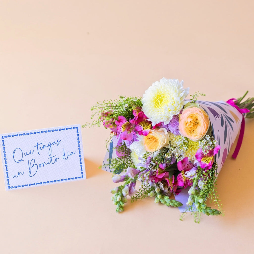 Pack La Costa de Colvin, que incluye un ramo de flores frescas de verano con rosas amarillas, crisantemos blancos y alstroemerias fucsias, envuelto en papel con motivos florales y acompañado de una tarjeta azul con el texto manuscrito "Que tengas un Bonito día" sobre fondo beige.