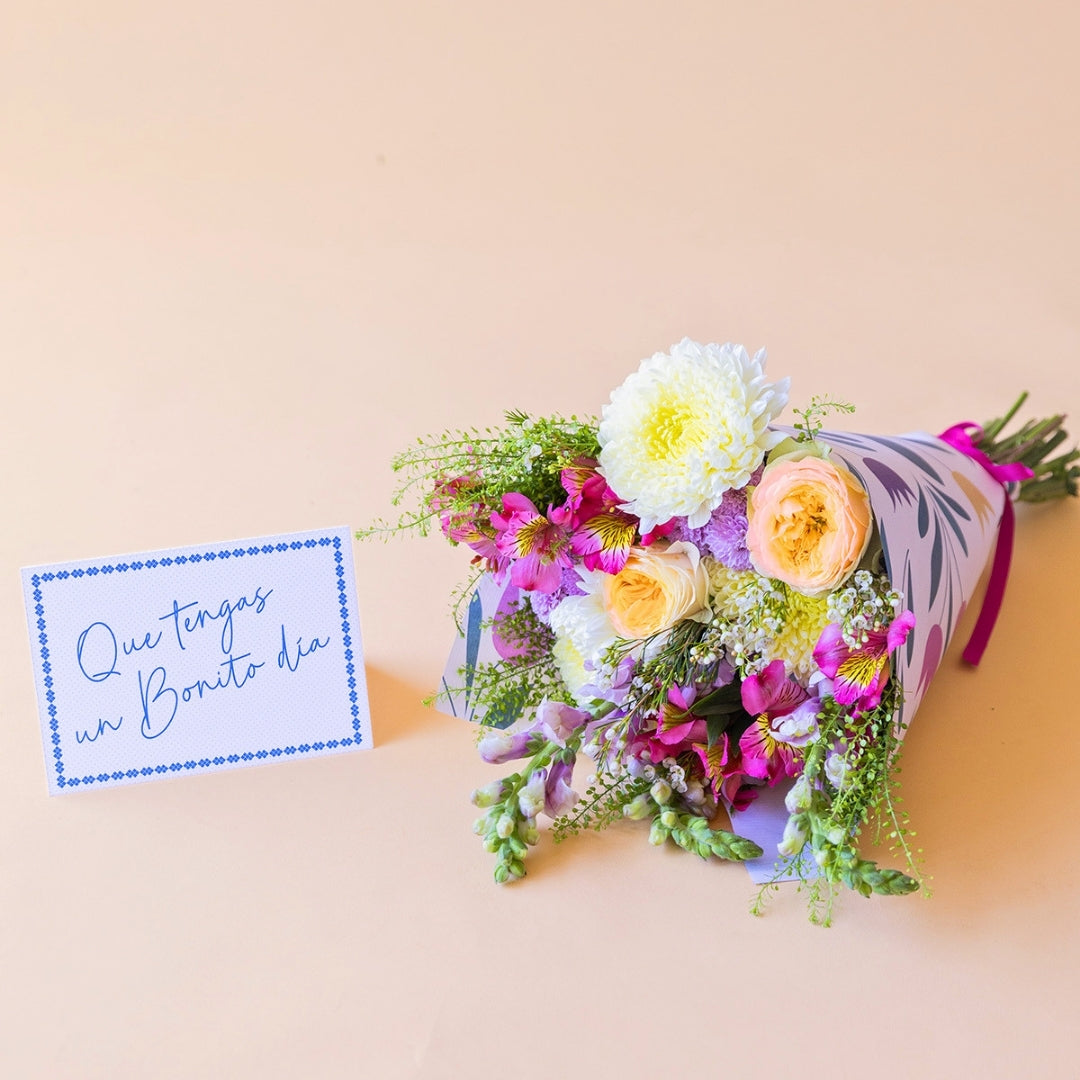 Pack La Costa de Colvin, que incluye un ramo de flores frescas de verano con rosas amarillas, crisantemos blancos y alstroemerias fucsias, envuelto en papel con motivos florales y acompañado de una tarjeta azul con el texto manuscrito "Que tengas un Bonito día" sobre fondo beige.
