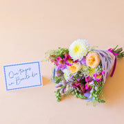 Pack La Costa de Colvin, que incluye un ramo de flores frescas de verano con rosas amarillas, crisantemos blancos y alstroemerias fucsias, envuelto en papel con motivos florales y acompañado de una tarjeta azul con el texto manuscrito "Que tengas un Bonito día" sobre fondo beige.
