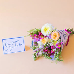 Pack La Costa de Colvin, que incluye un ramo de flores frescas de verano con rosas amarillas, crisantemos blancos y alstroemerias fucsias, envuelto en papel con motivos florales y acompañado de una tarjeta azul con el texto manuscrito "Que tengas un Bonito día" sobre fondo beige.
