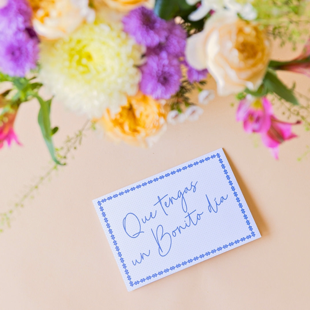 Detalle del pack La Costa de Colvin, que incluye un ramo de flores frescas de verano con rosas amarillas, crisantemos blancos y alstroemerias fucsias, acompañado de una tarjeta azul con el texto manuscrito "Que tengas un Bonito día" sobre fondo beige.