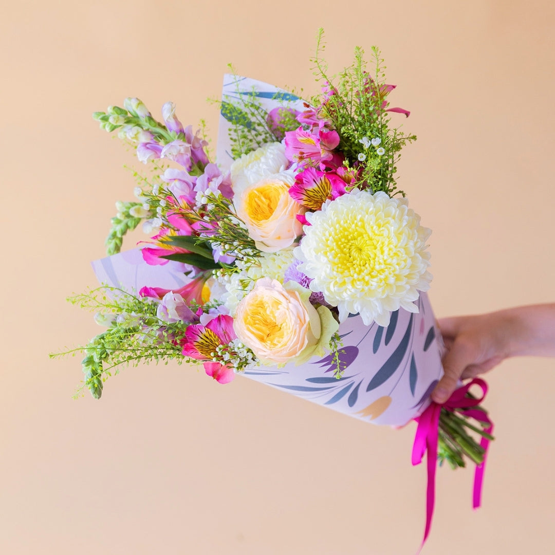Pack La Costa de Colvin, que incluye un ramo de flores frescas de verano con rosas amarillas, crisantemos blancos y alstroemerias fucsias, envuelto en papel con motivos florales.