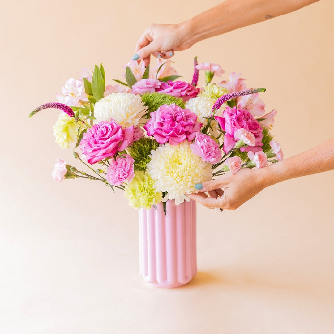 Manos arreglando el ramo La Dolce Vita de Colvin en jarrón acanalado rosa con rosas, crisantemos blancos y flores silvestres, sobre fondo melocotón